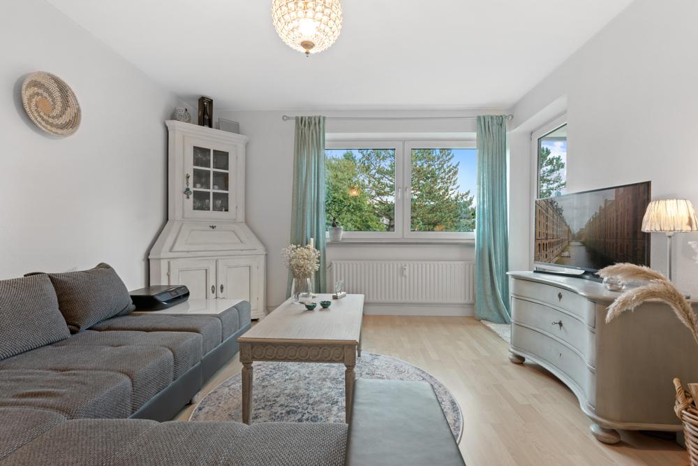 Helles Wohnzimmer mit grauem Sofa, weißem Schrank, Couchtisch und großem Fenster mit Vorhängen.