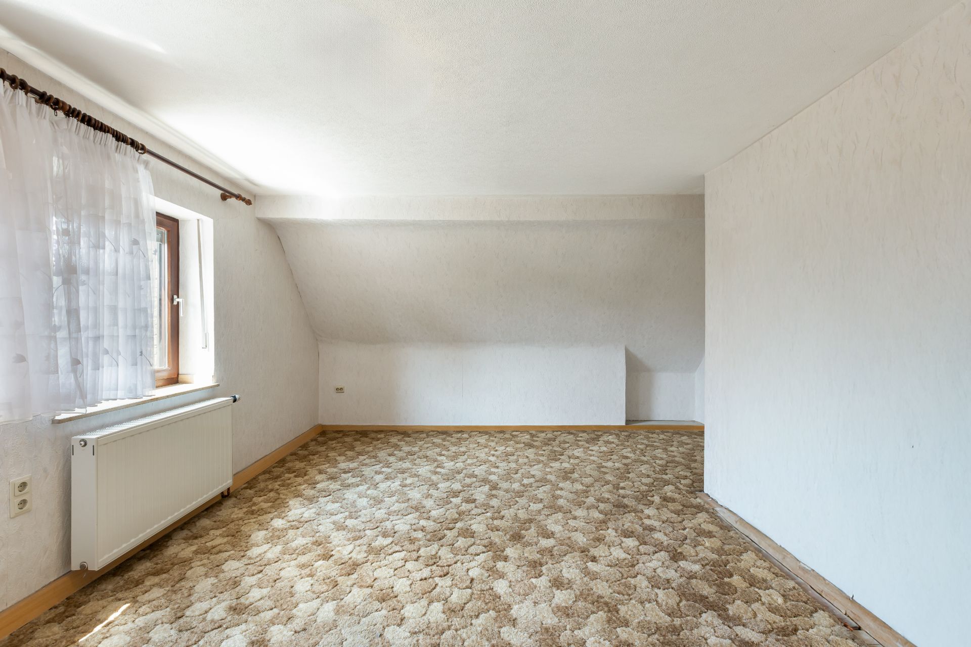 Leeres Zimmer mit gemustertem Teppich, weißem Heizkörper und Fenster mit Vorhang.