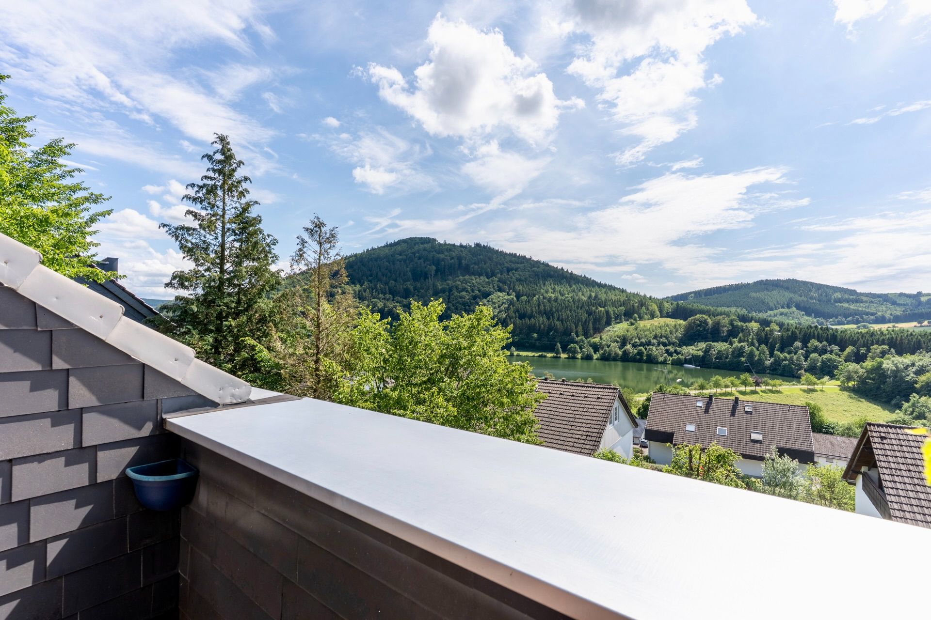 Blick vom Balkon auf grüne Hügel, Wald und einen kleinen See, Häuser im Vordergrund.