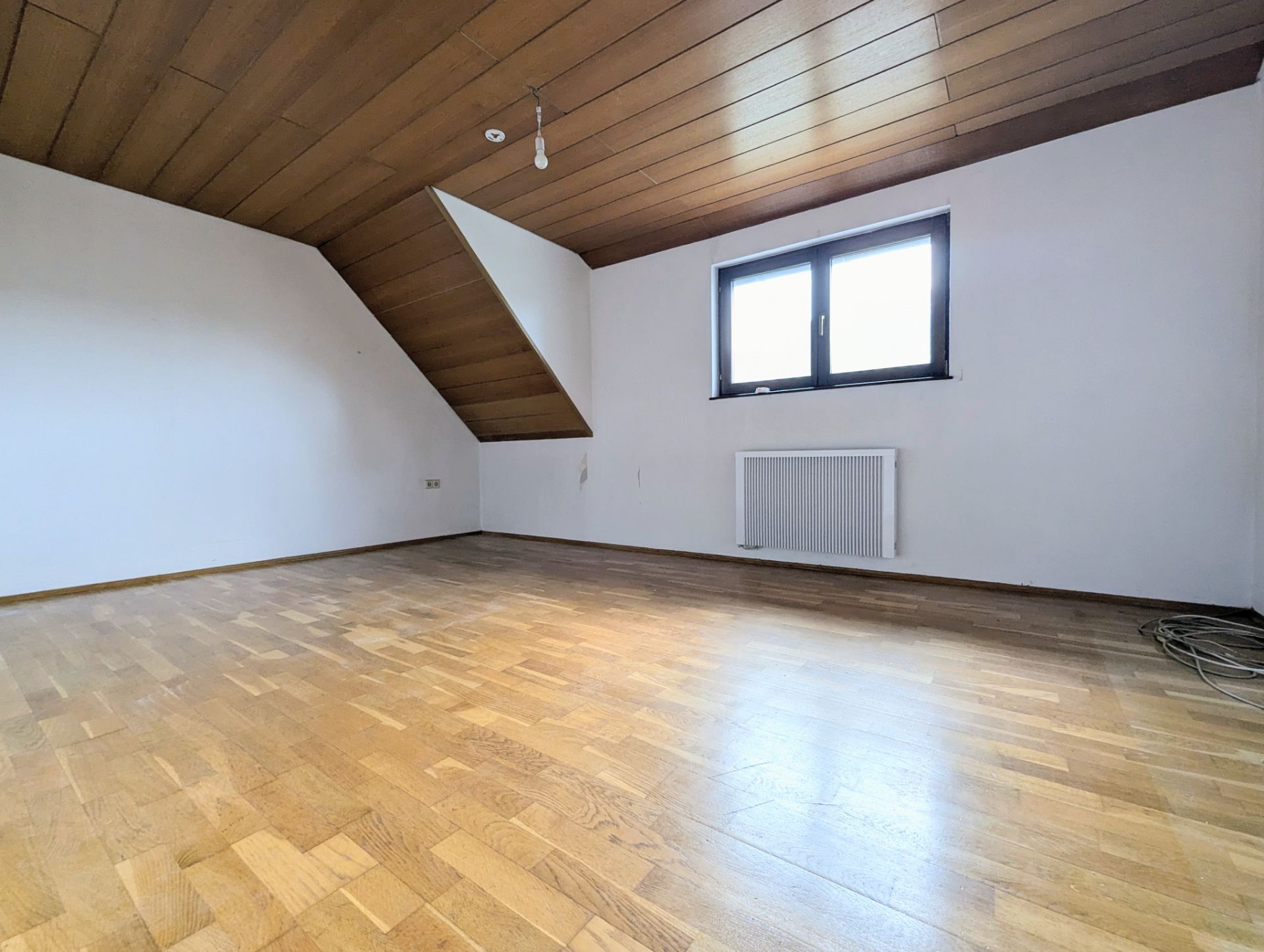 Leeres Zimmer mit Holzdecke, Holzfußboden, Fenster und Heizkörper an der Wand.