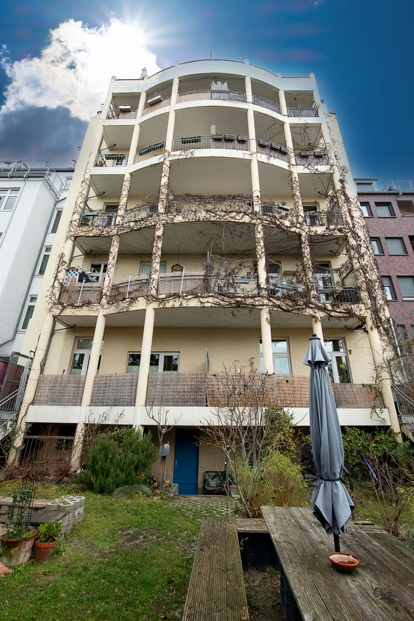Mehrstöckiges Wohnhaus mit Balkonen und Kletterpflanzen, im Vordergrund Garten mit Bank und Sonnenschirm.