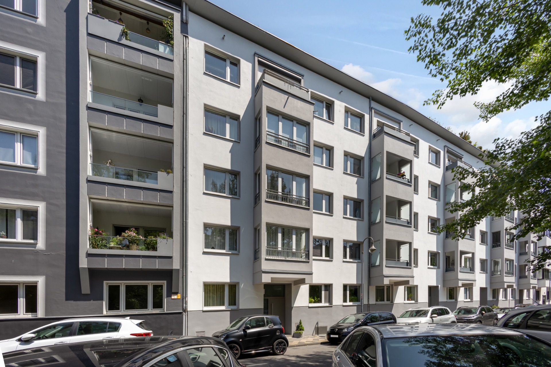 Mehrfamilienhaus mit Balkonen und parkenden Autos davor an einer Straße.