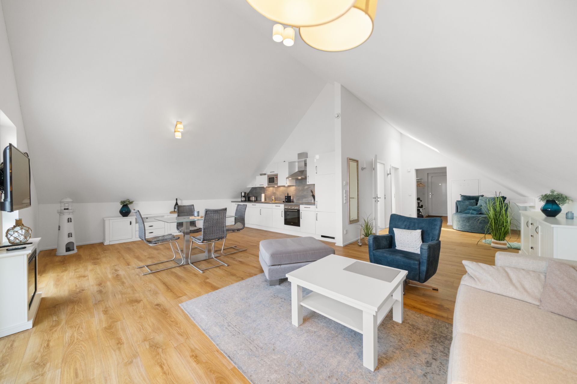Helles Wohnzimmer mit Küchenzeile, Esstisch, Sofa und Sesseln unter Dachschrägen.