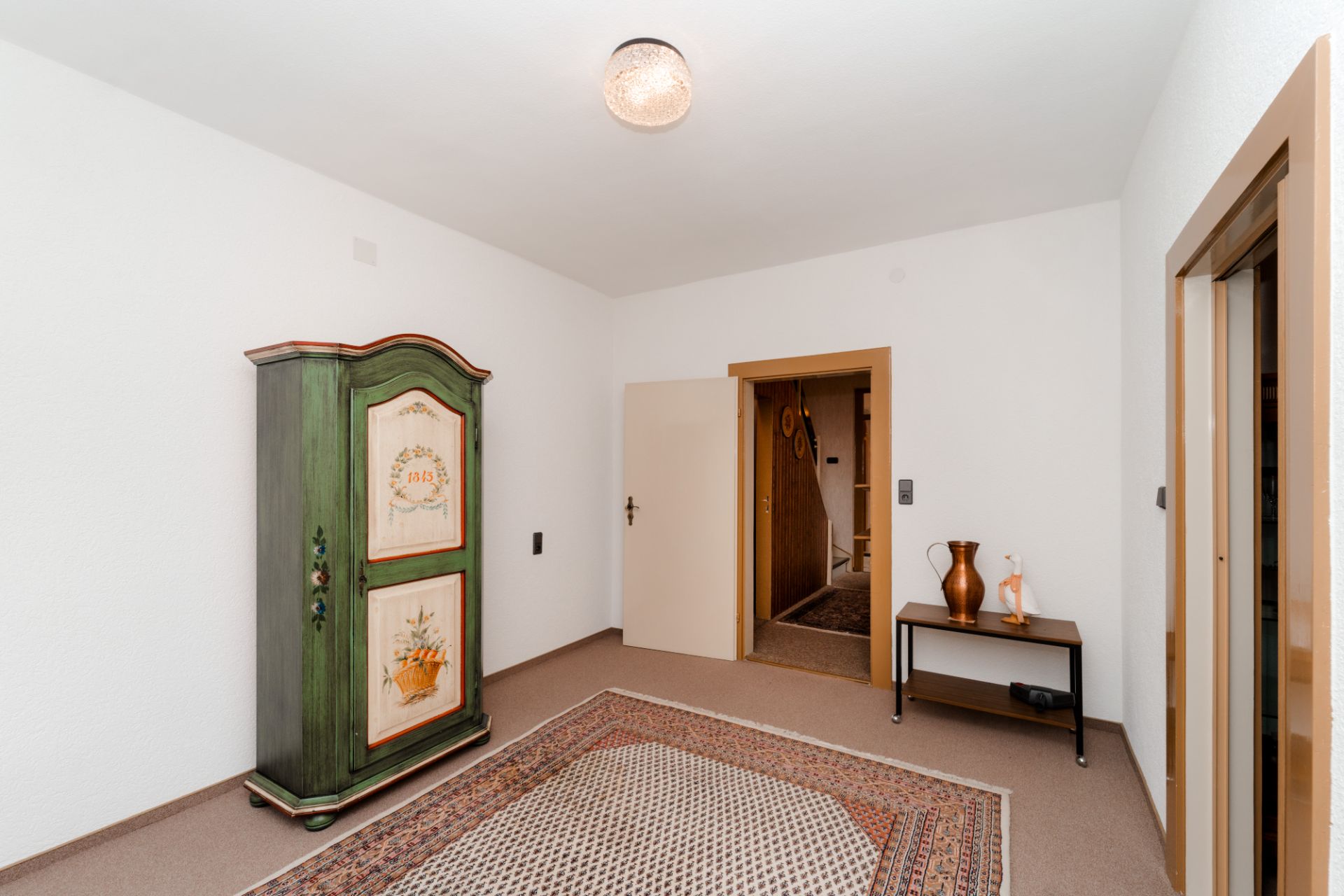 Zimmer mit grünem Schrank, Teppich und kleinem Tisch mit Vasen an weißer Wand.