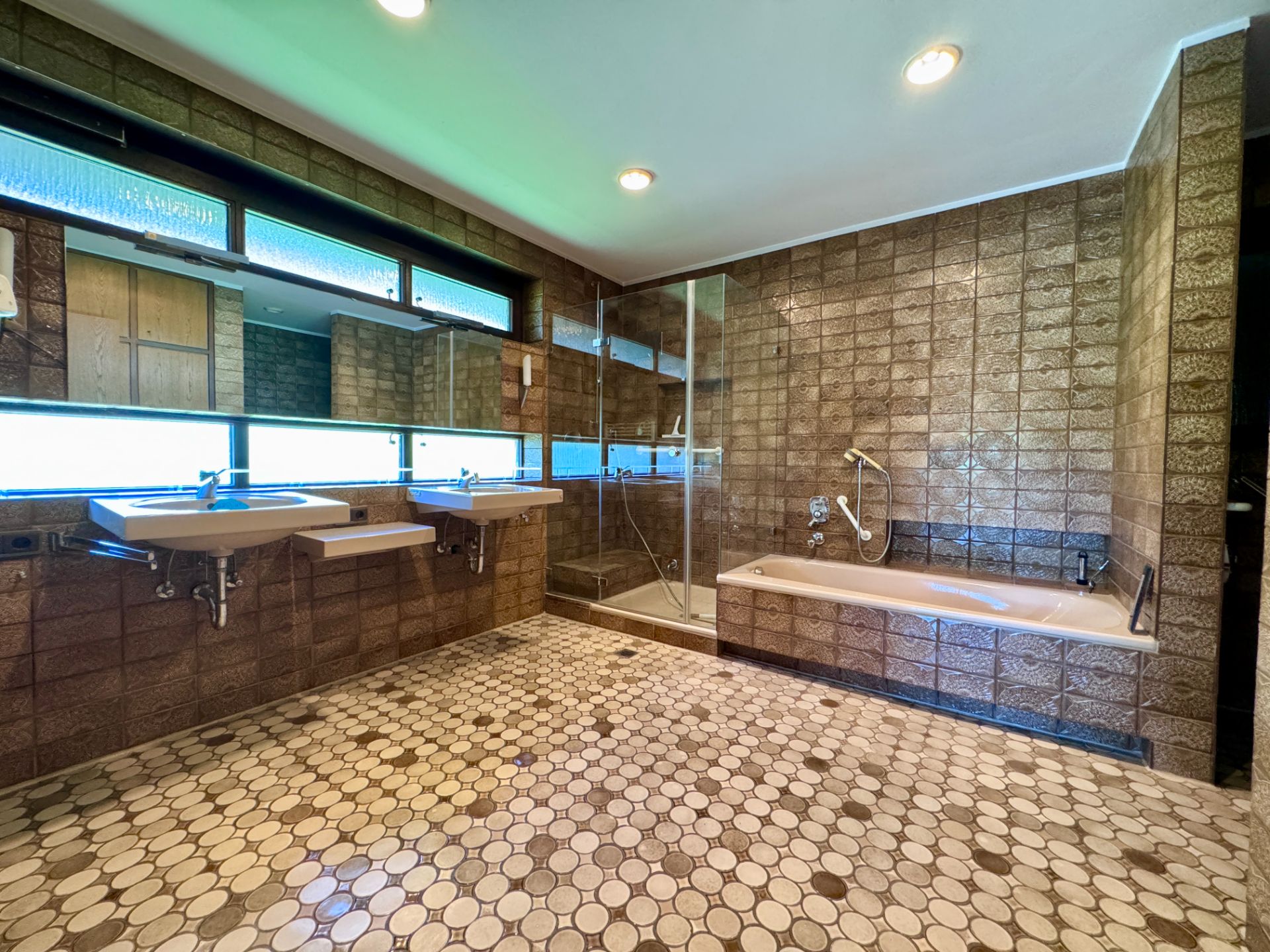 Großes Badezimmer mit zwei Waschbecken, Dusche und Badewanne, braune Fliesen und gemusterter Boden.