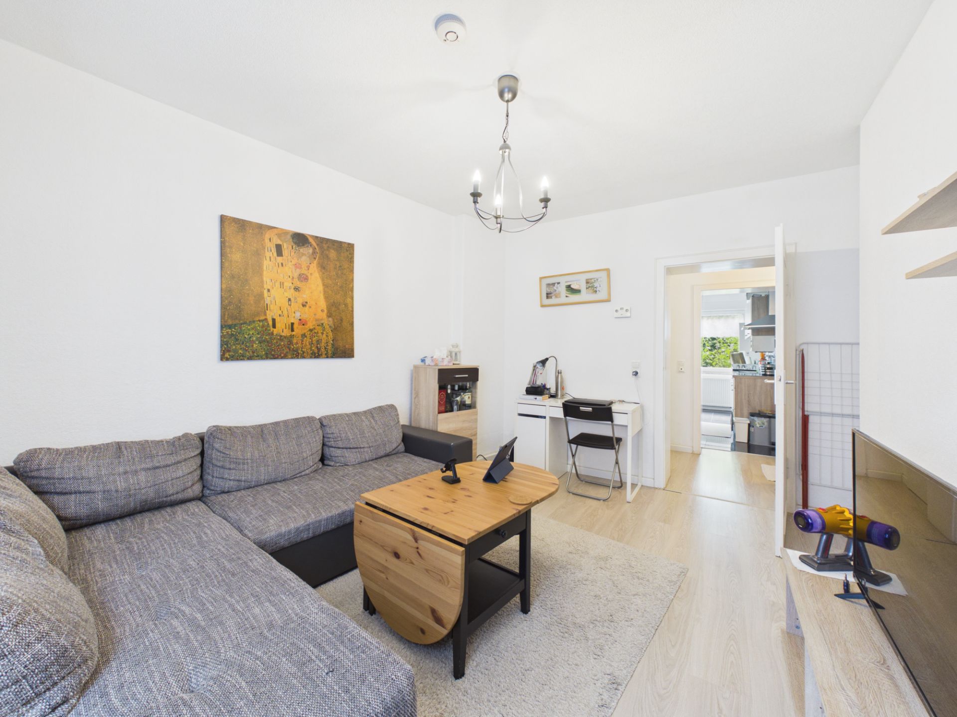 Wohnzimmer mit grauem Ecksofa, Holztisch, Schreibtisch und Wandbild, TV rechts.