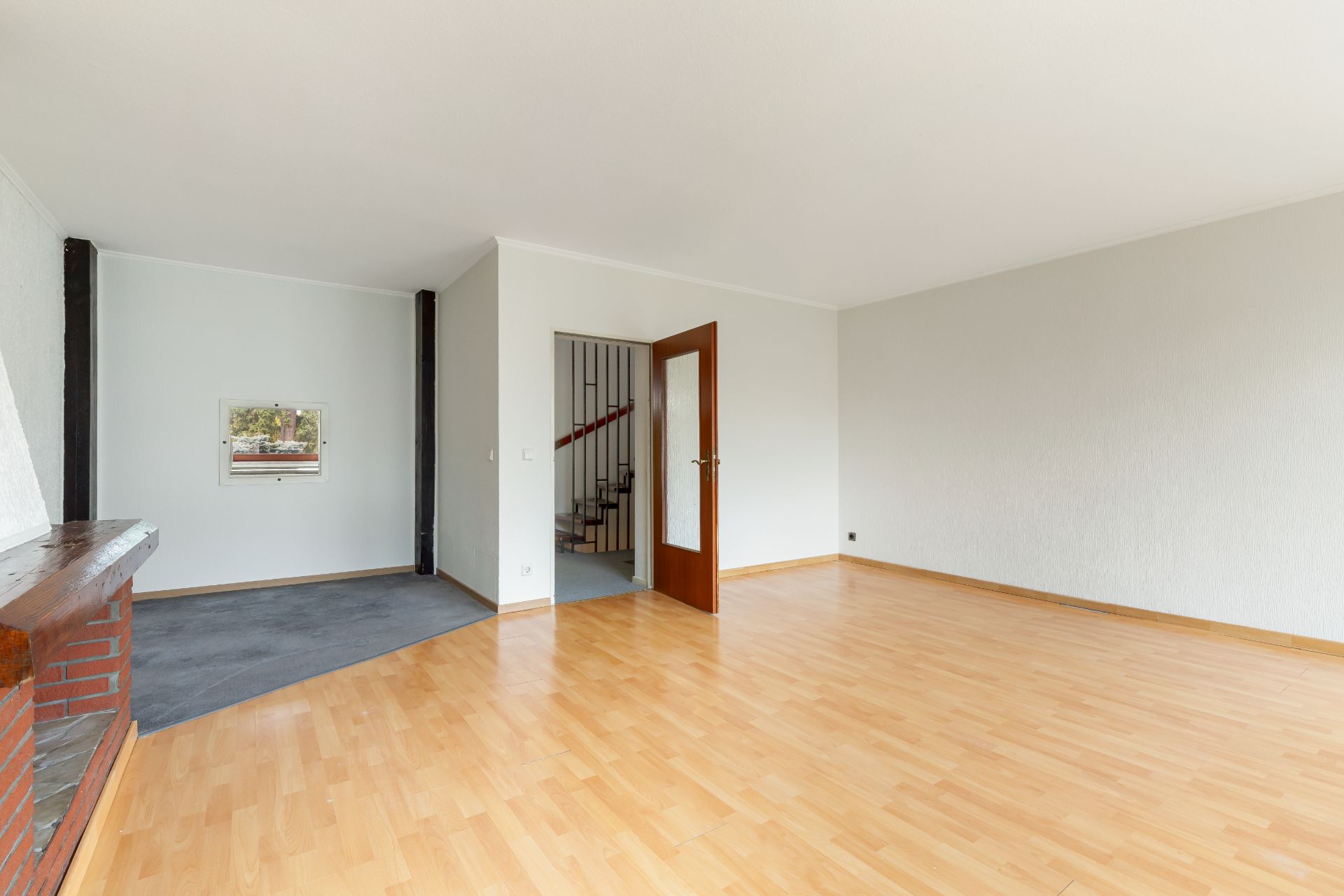 Leeres Wohnzimmer mit Parkettboden, Kamin und Blick auf eine Treppe durch eine offene Tür.