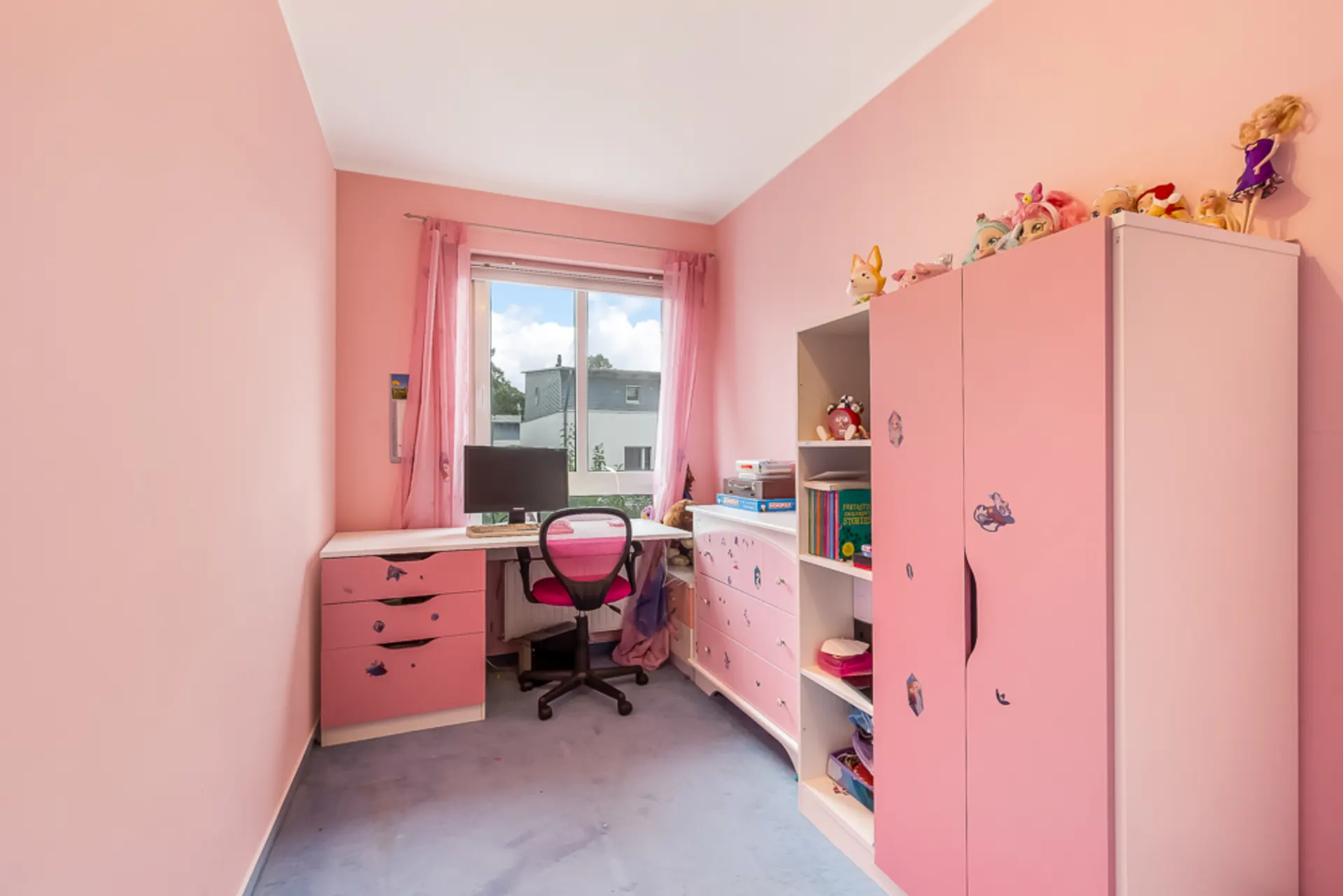 Kinderzimmer mit rosa Wänden, Schreibtisch, Stuhl, Schrank und Regal, Spielzeug darauf.