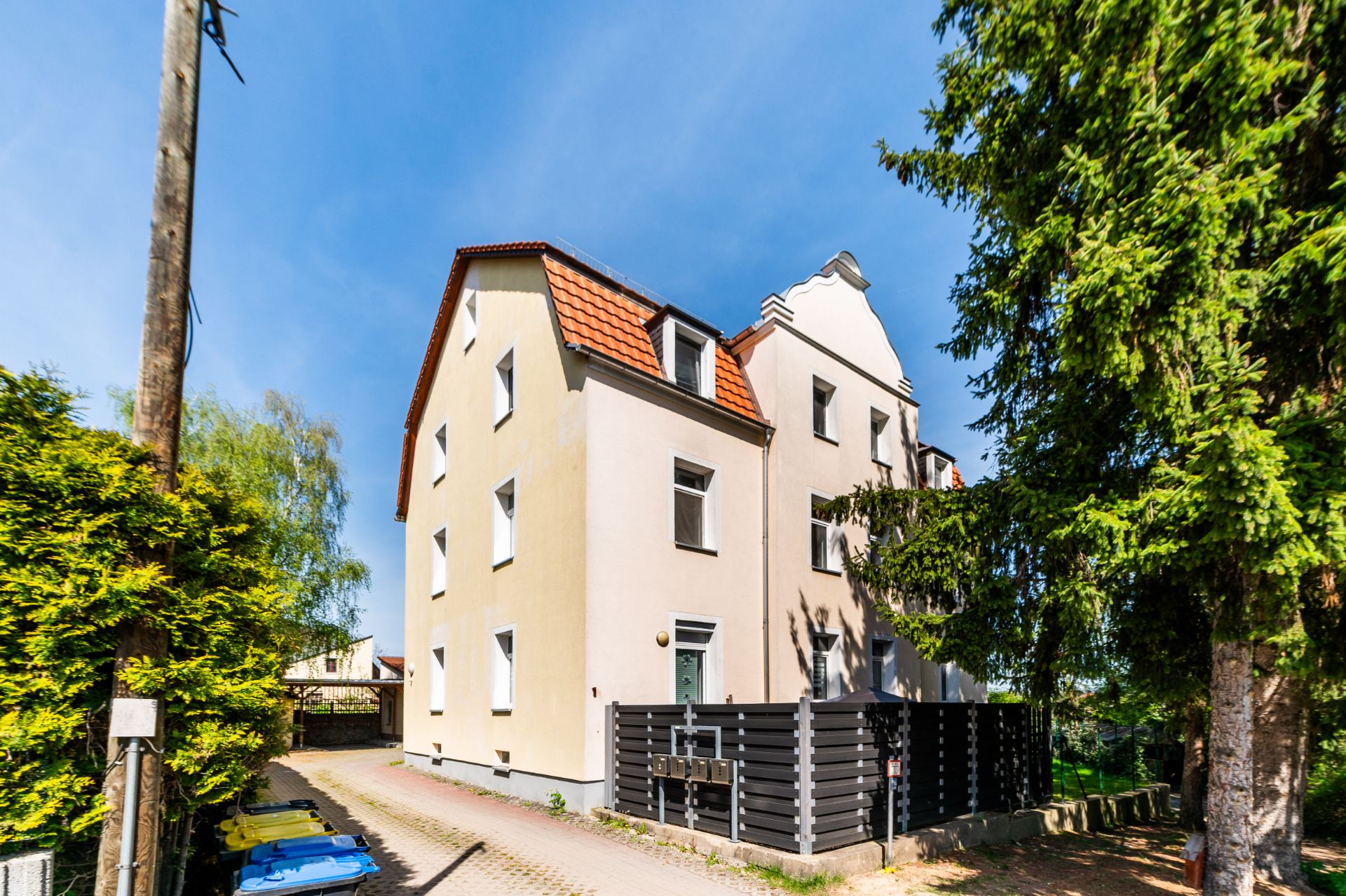 Helles, dreistöckiges Wohnhaus mit roten Dachziegeln und seitlichem Zaun, Bäume am Rand.