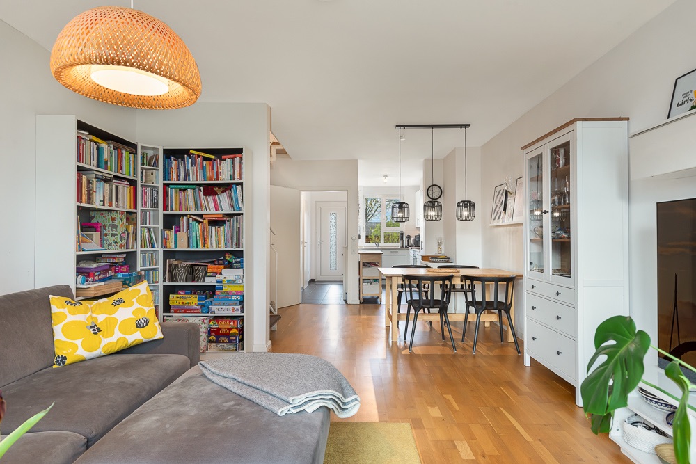 Helles Wohnzimmer mit Sofa, Bücherregal, Esstisch und offener Küche auf Holzboden.