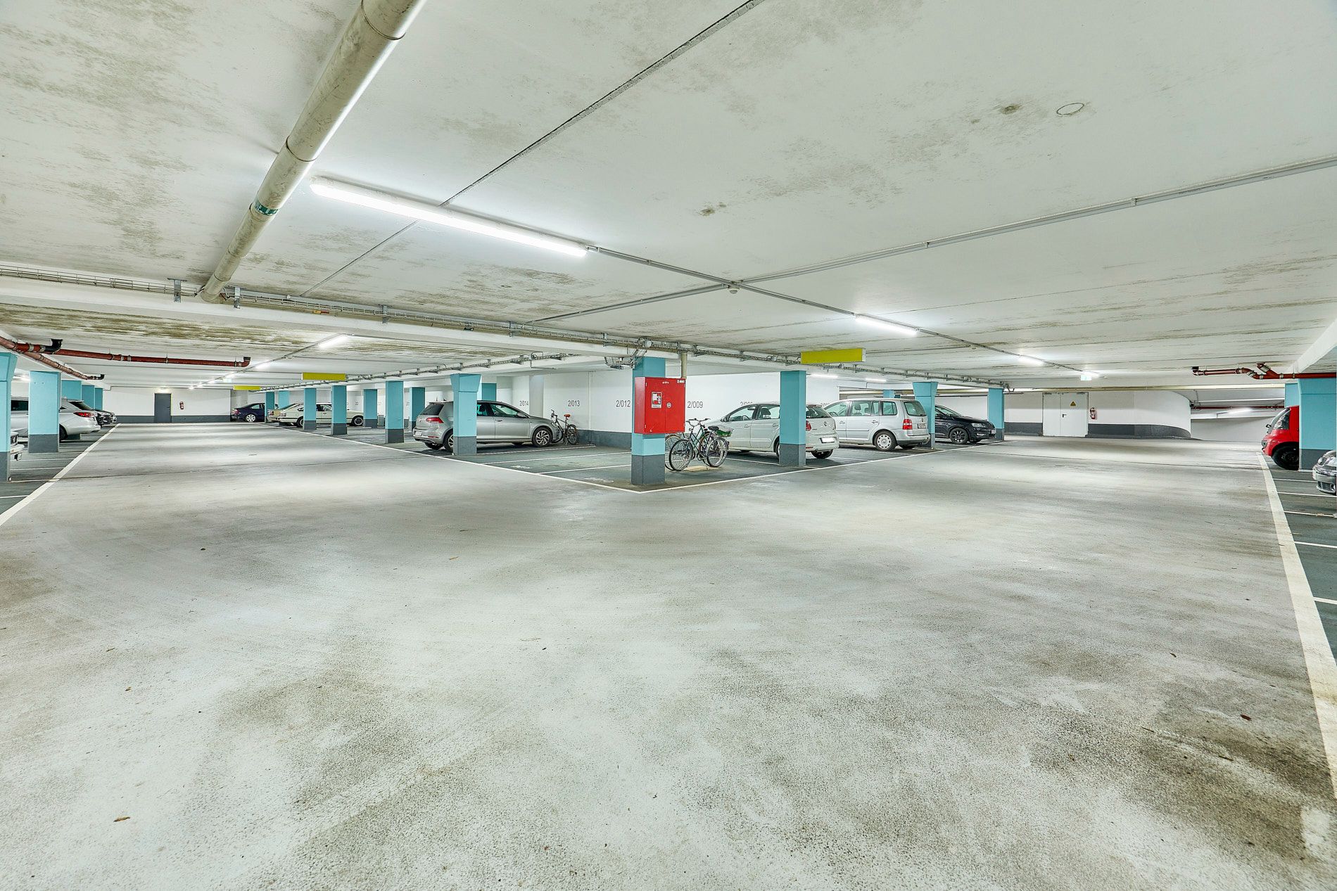 Helle Tiefgarage mit geparkten Autos, Fahrrädern und markierten Stellplätzen.