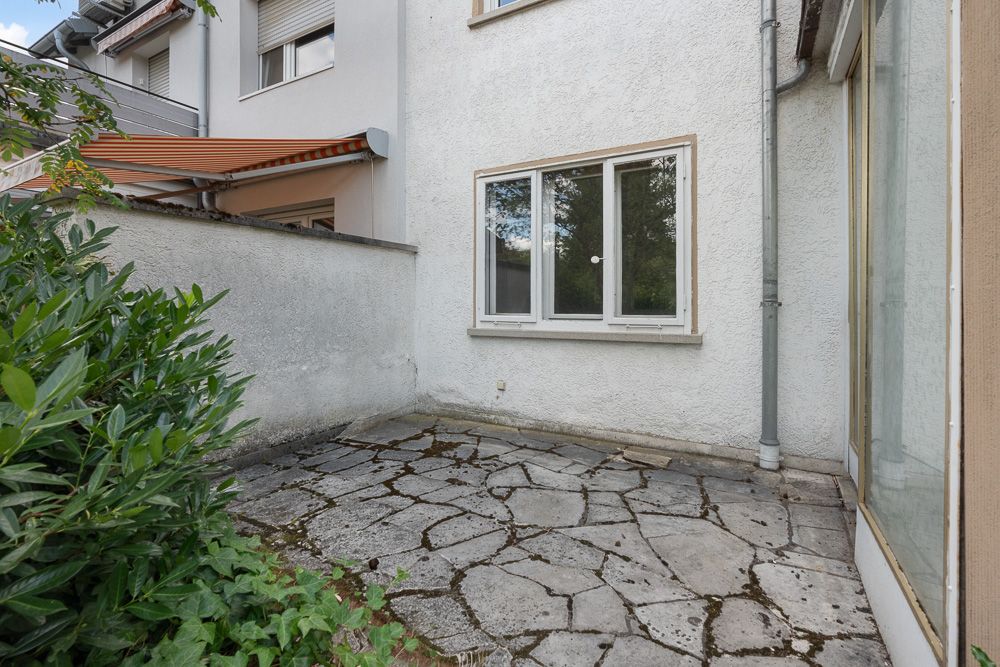 Kleine, steingepflasterte Terrasse mit einer weißen Hauswand und Fenstern.