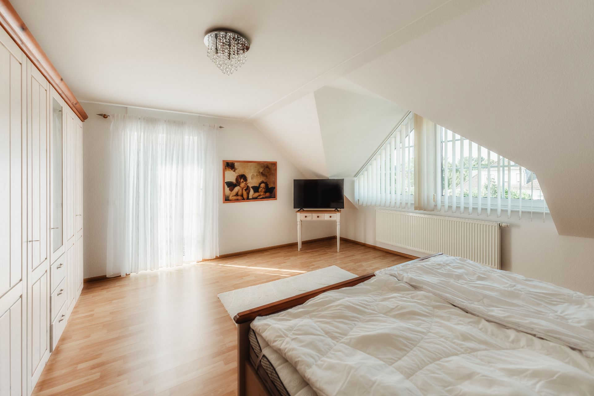 Helles Schlafzimmer mit Holzboden, großem Schrank, Bett, TV auf einem Tisch und großem Fenster.