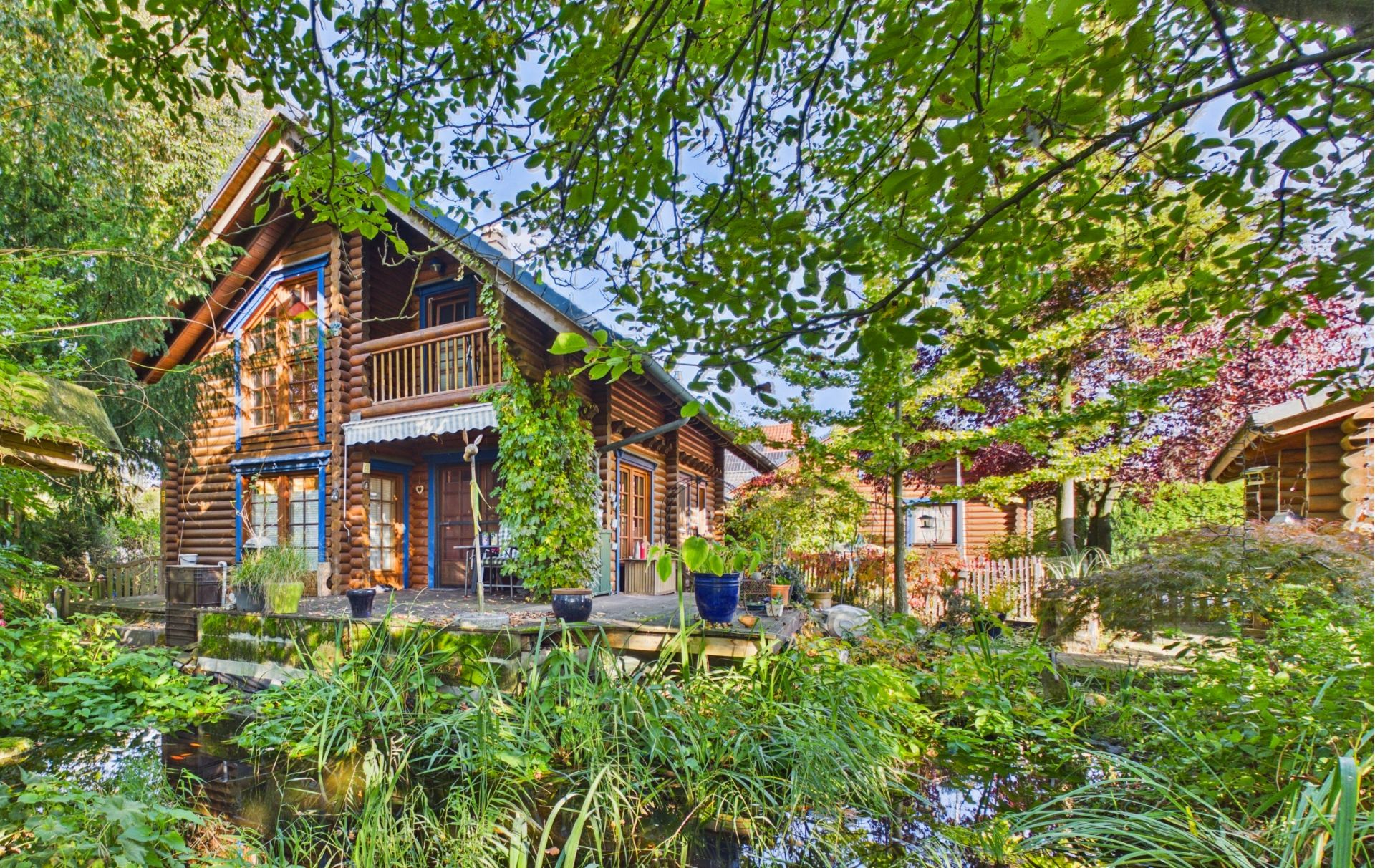 Blockhaus mit Balkon und Terrasse, umgeben von Garten und Teich, viele grüne Pflanzen im Vordergrund.