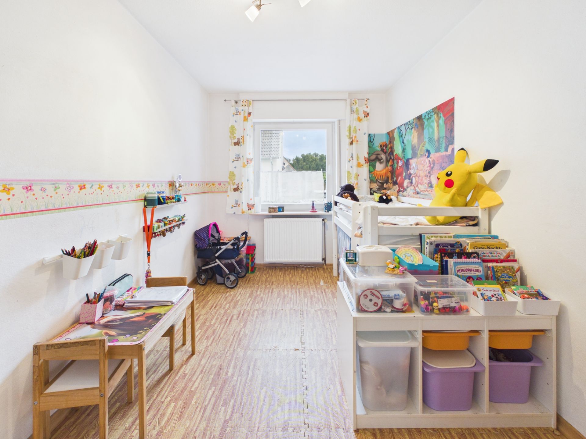 Helles Kinderzimmer mit Spielsachen, Regal, Schreibtisch und Fenster am Ende des Raumes.