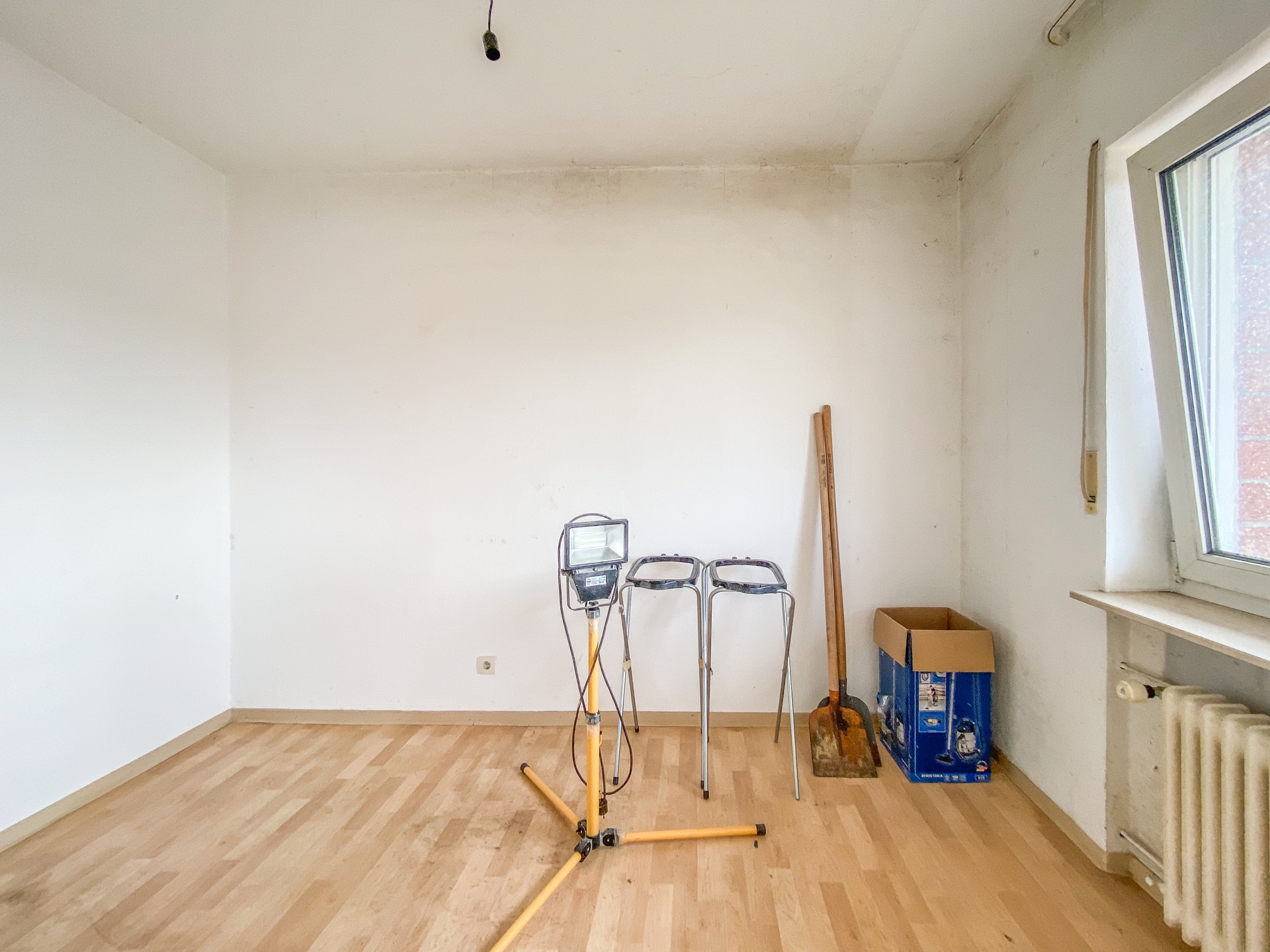 Leeres Zimmer mit Holzfußboden, Scheinwerfer, zwei Hockern, Karton, Werkzeug und Fenster.