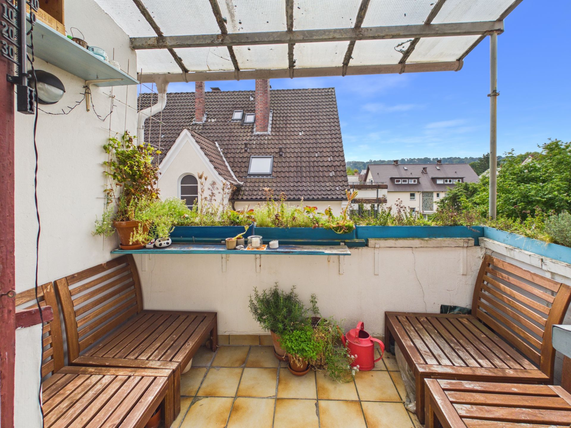 Kleine überdachte Terrasse mit Holzbänken, Pflanzen und Blick auf Nachbarhäuser.
