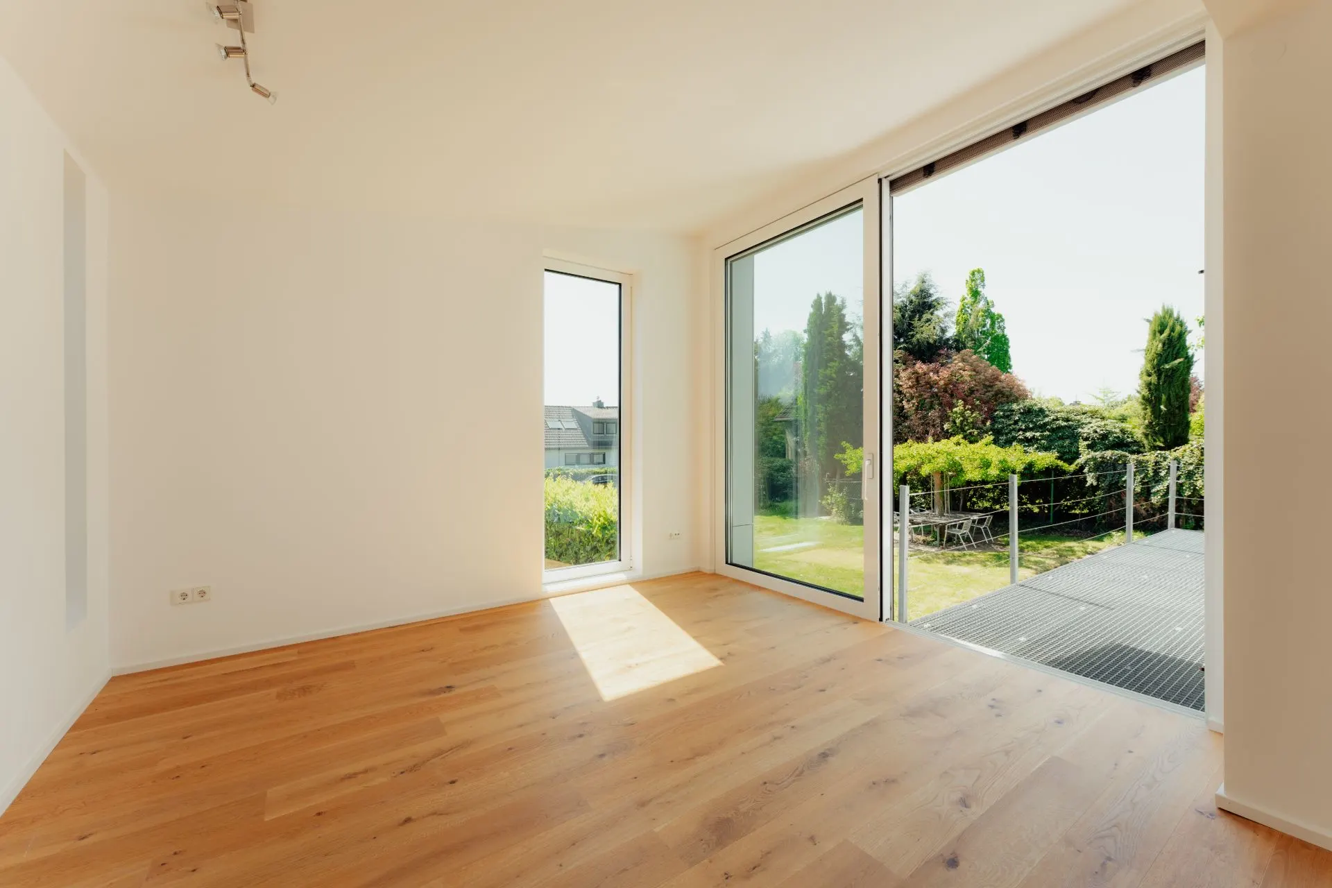Leeres Zimmer mit Holzboden und großen Fenstern mit Blick auf einen Garten.