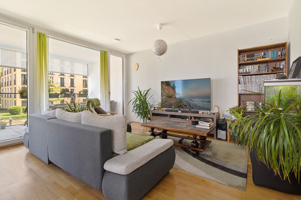 Helles Wohnzimmer mit Sofa, Couchtisch, Fernseher, Bücherregal und vielen Pflanzen.
