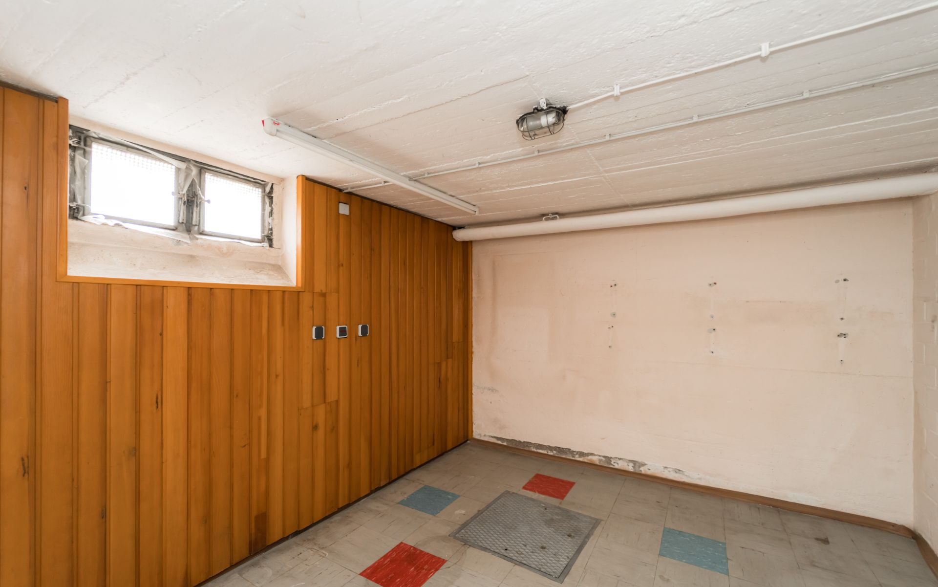 Kleiner Raum mit Holzwand und kleinem Fenster, bunte Fliesen auf dem Boden.