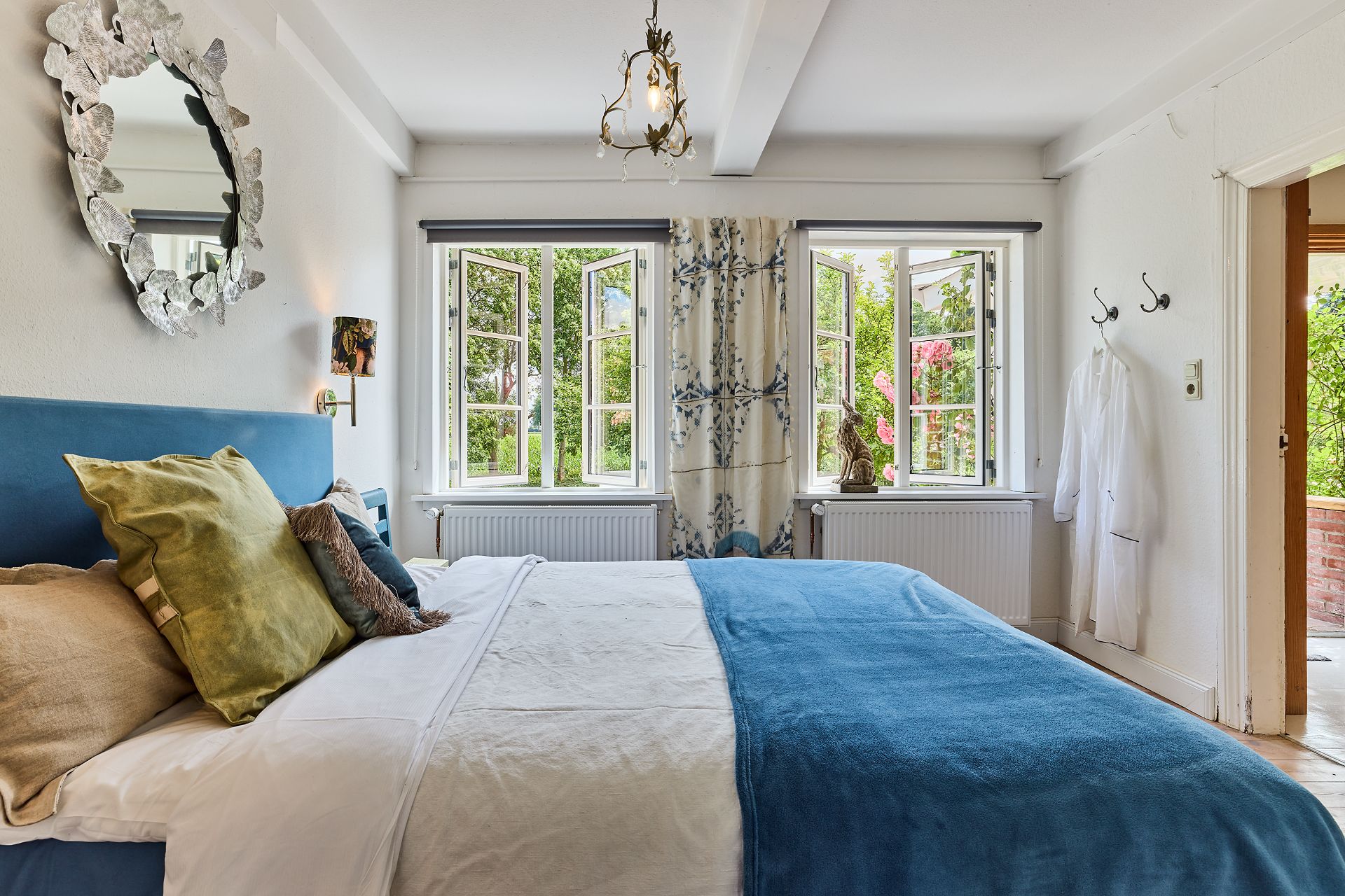 Helles Schlafzimmer mit Doppelbett, offenen Fenstern und Spiegel an der Wand.