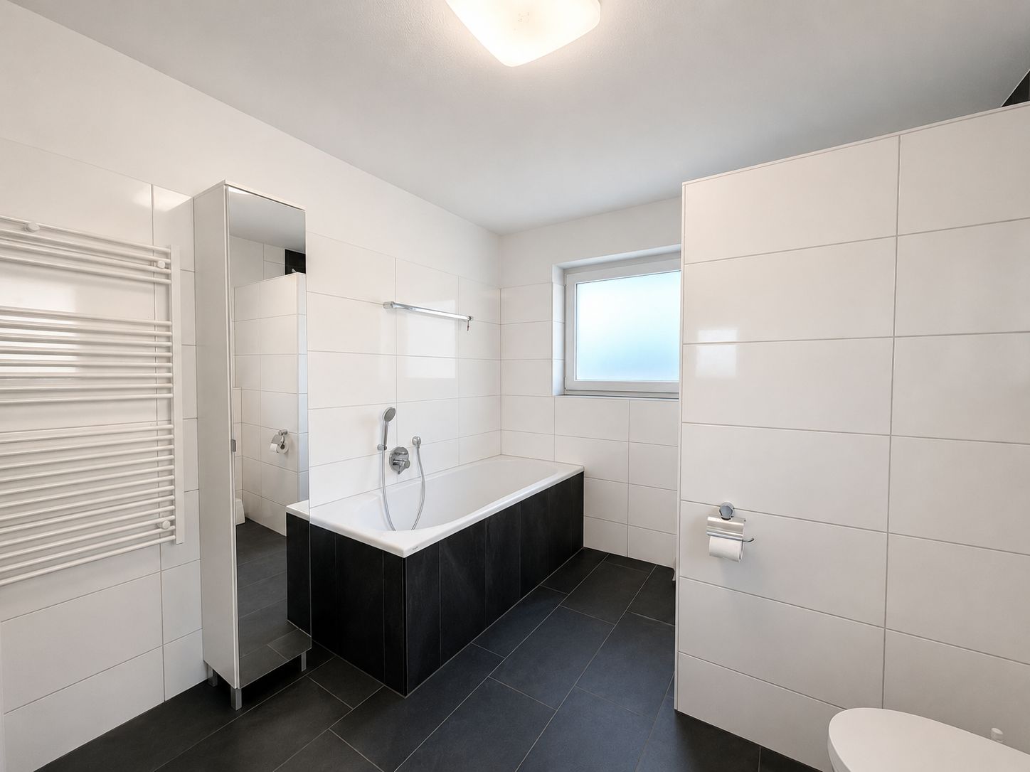 Modernes Badezimmer mit Badewanne, Spiegel, Handtuchheizkörper und Fenster.