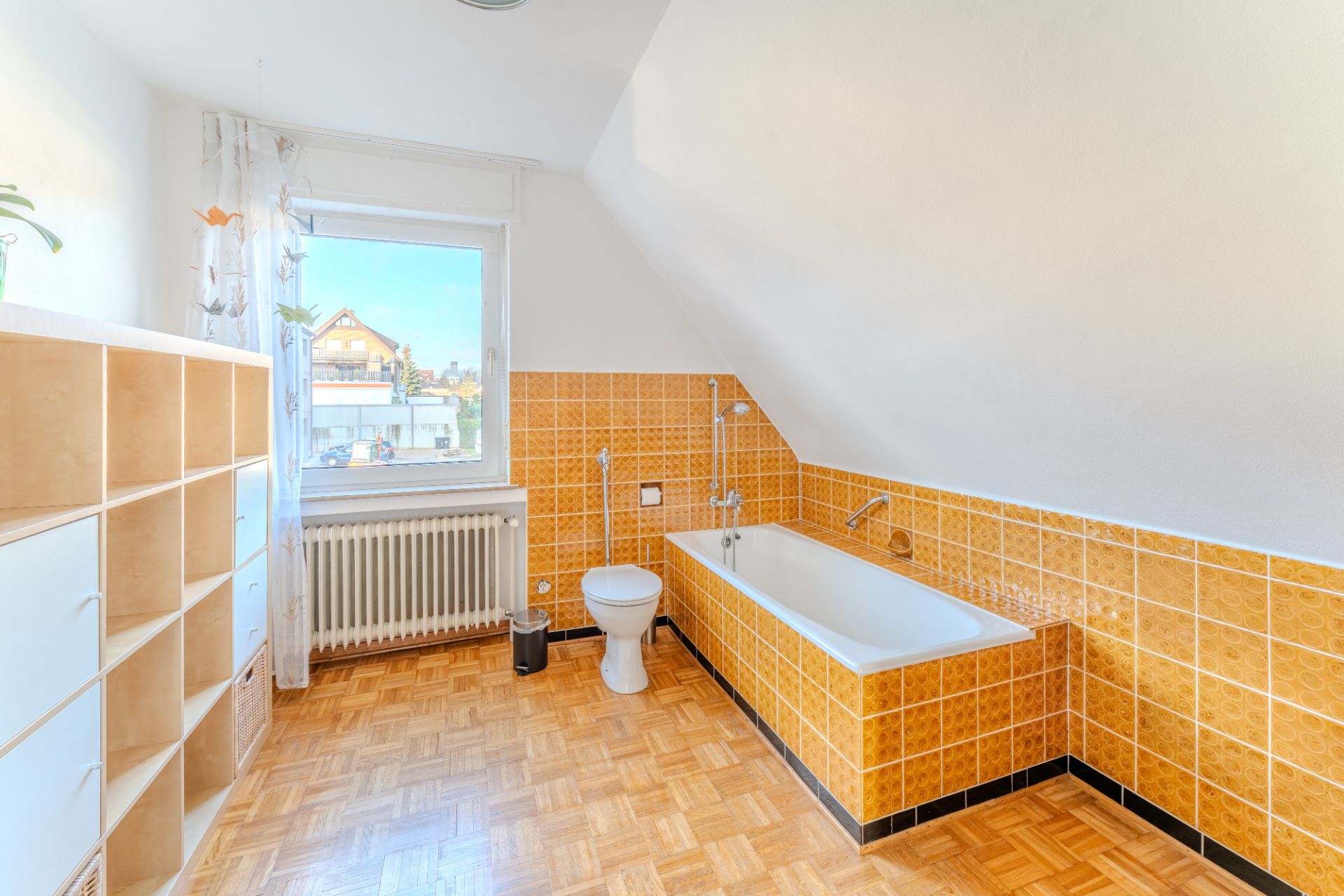 Helles Badezimmer mit Badewanne, Toilette, gelben Fliesen und Regal auf Parkettboden.