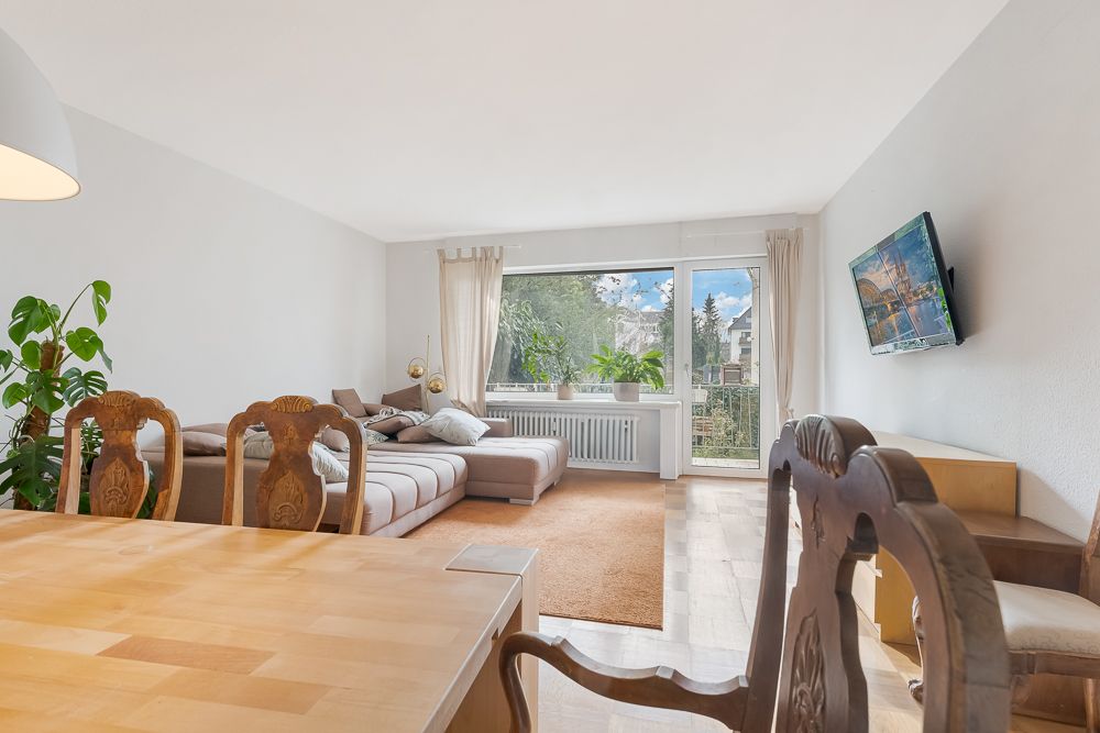 Helles Wohnzimmer mit großer Couch, Esstisch, Pflanzen, Fenster und TV an der Wand.