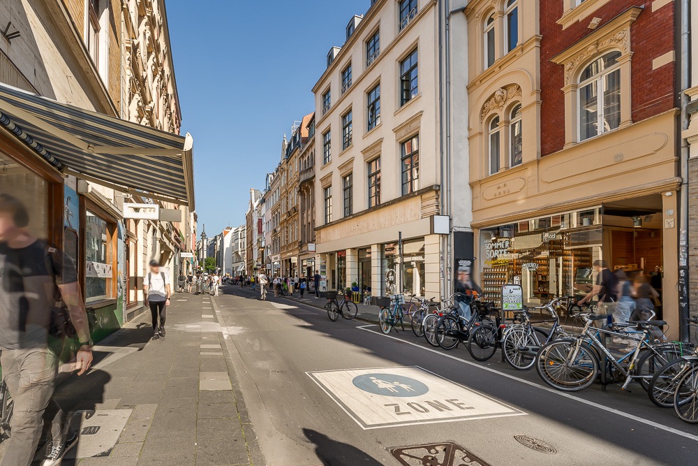 Belebte Straße mit Gehweg, Fahrrädern und mehreren mehrstöckigen Wohngebäuden bei sonnigem Wetter.