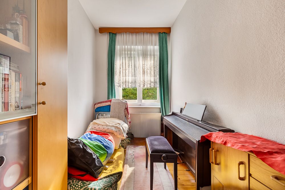 Kleines Zimmer mit Einzelbett, E-Piano, Stuhl und Fenster mit grünen Vorhängen.