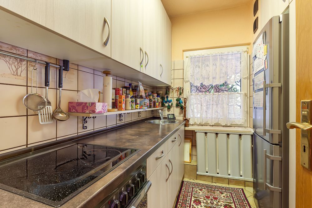 Schmale Küche mit Herd, Kühlschrank, Hängeschränken und Fenster mit Gardine.