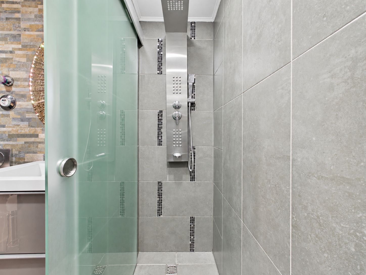Moderne Dusche mit grauen Fliesen und Glas-Schiebetür, daneben Teil einer Badewanne sichtbar.
