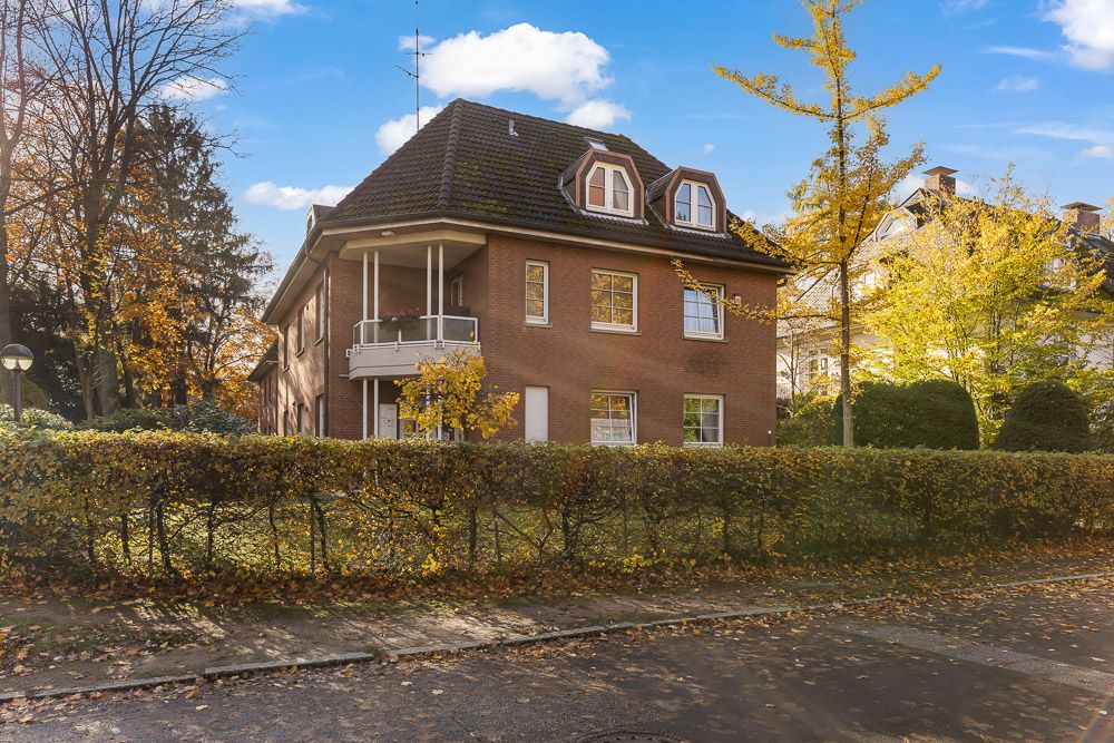 Backsteinhaus mit schwarzem Dach, Balkon und Garten, von Bäumen und Hecken umgeben.