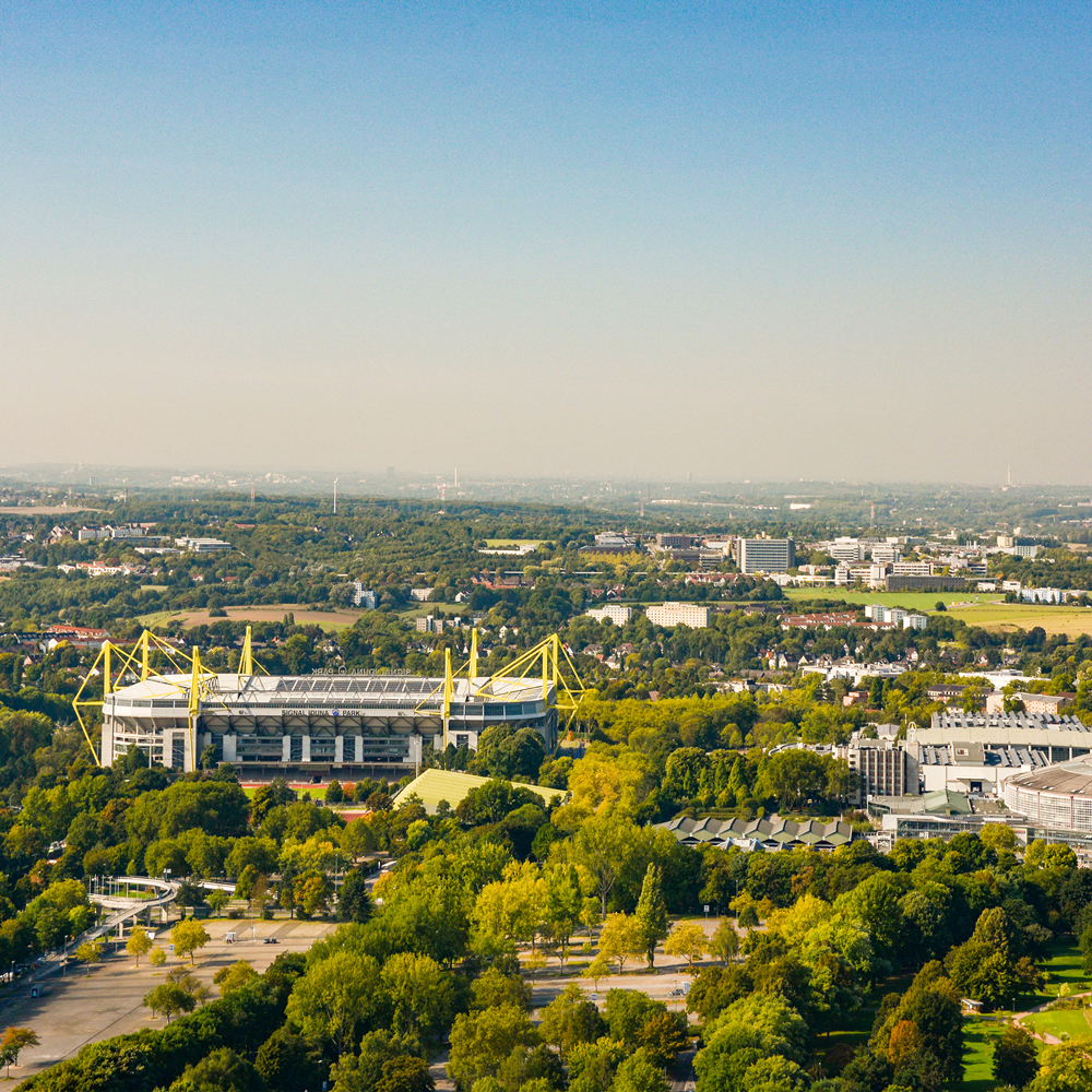 Luftaufnahme eines großen Stadions mit gelben Elementen, umgeben von vielen Bäumen und Gebäuden.