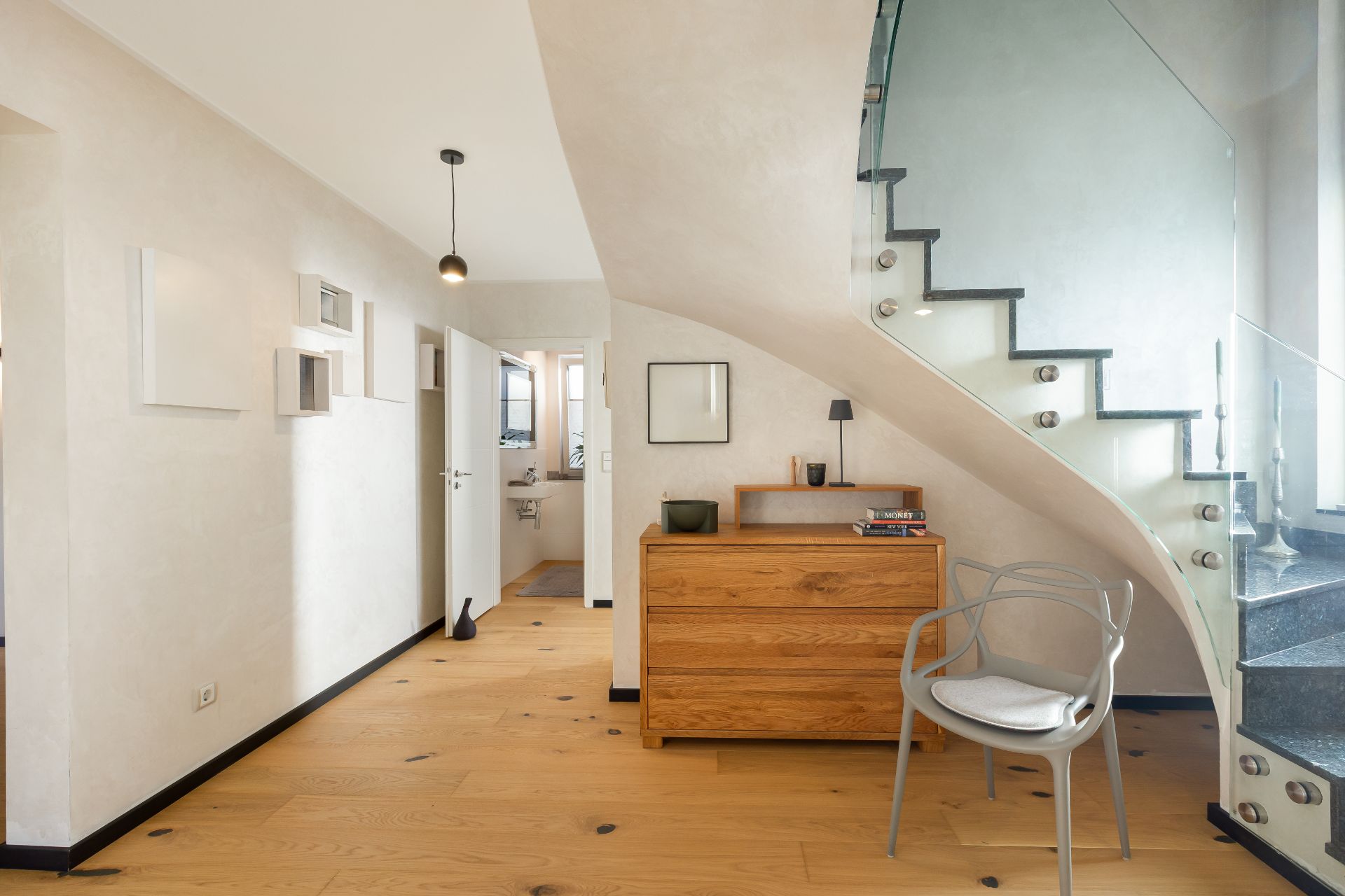 Heller Flur mit Holzkommode, Stuhl und Treppe mit Glasgeländer.