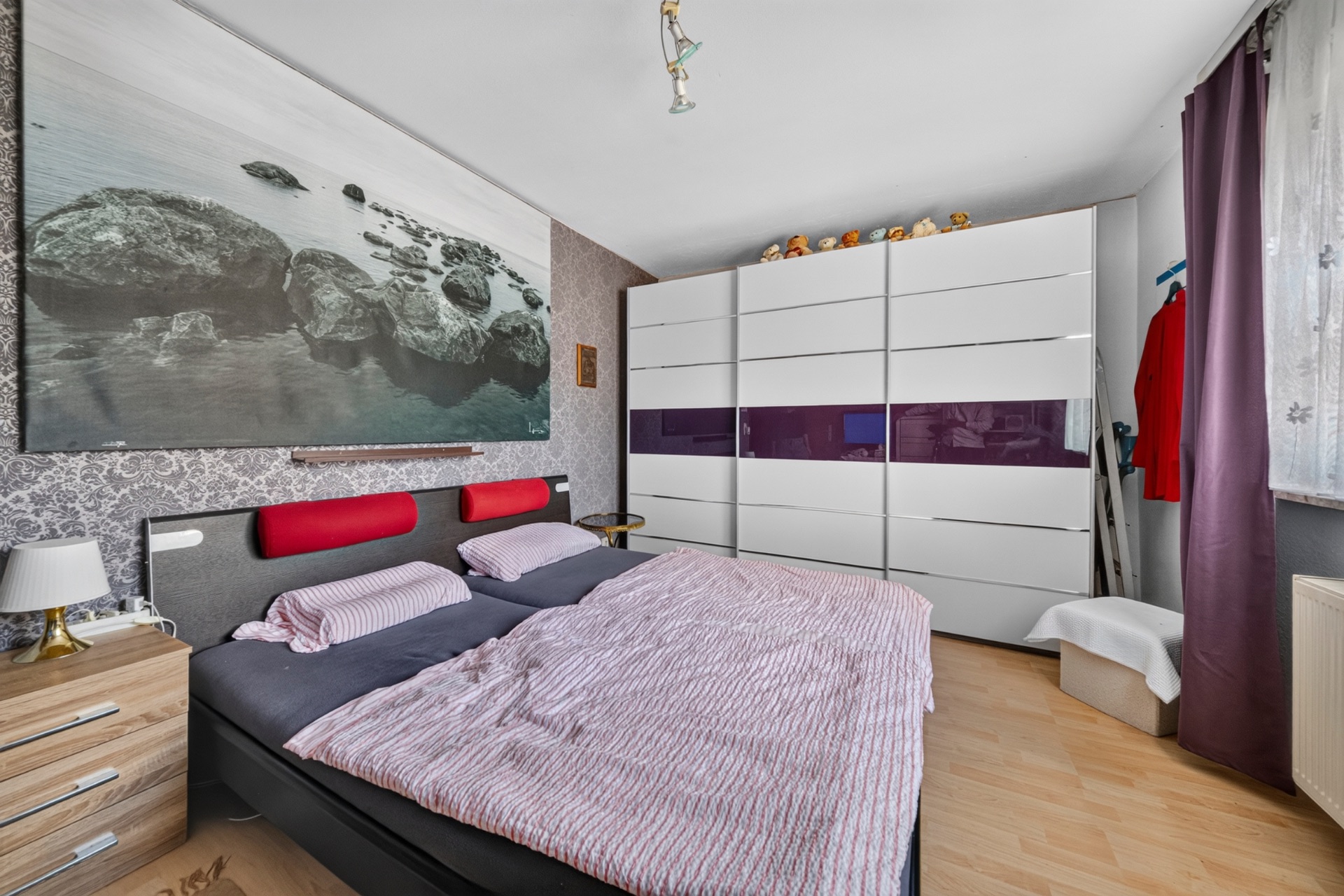 Schlafzimmer mit Doppelbett, großem Kleiderschrank und Fenster mit Vorhängen.