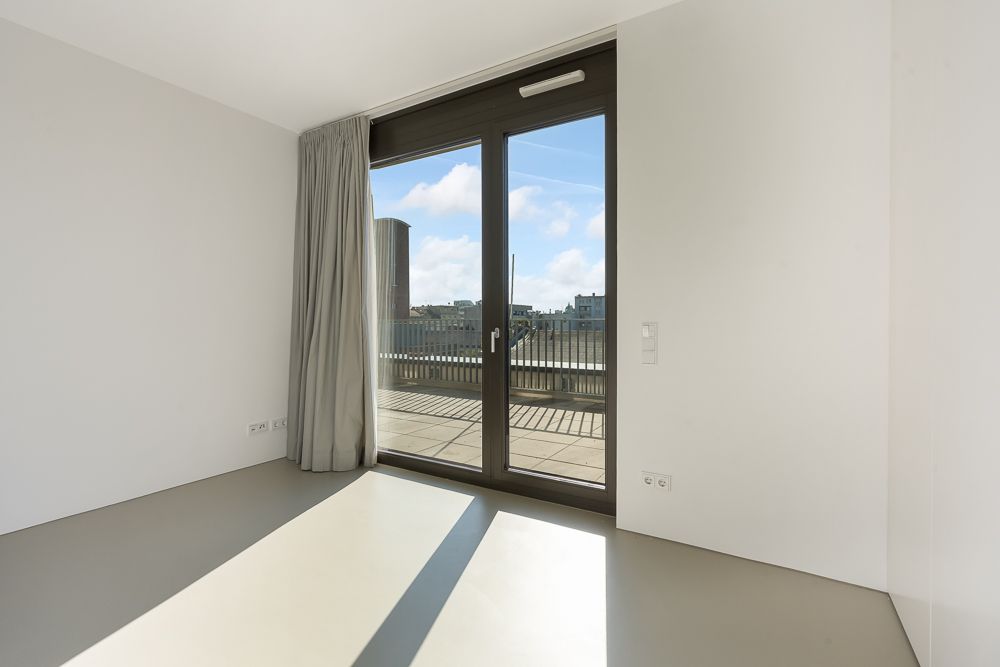 Helles Zimmer mit großen Fenstern und Zugang zu einem Balkon, Aussicht auf Stadtgebäude.
