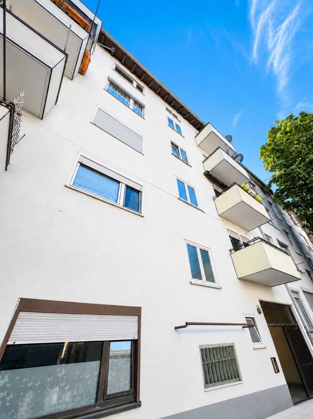Mehrstöckiges Wohnhaus mit weißen Fassaden, Fenstern und mehreren Balkonen, Baum am rechten Rand.
