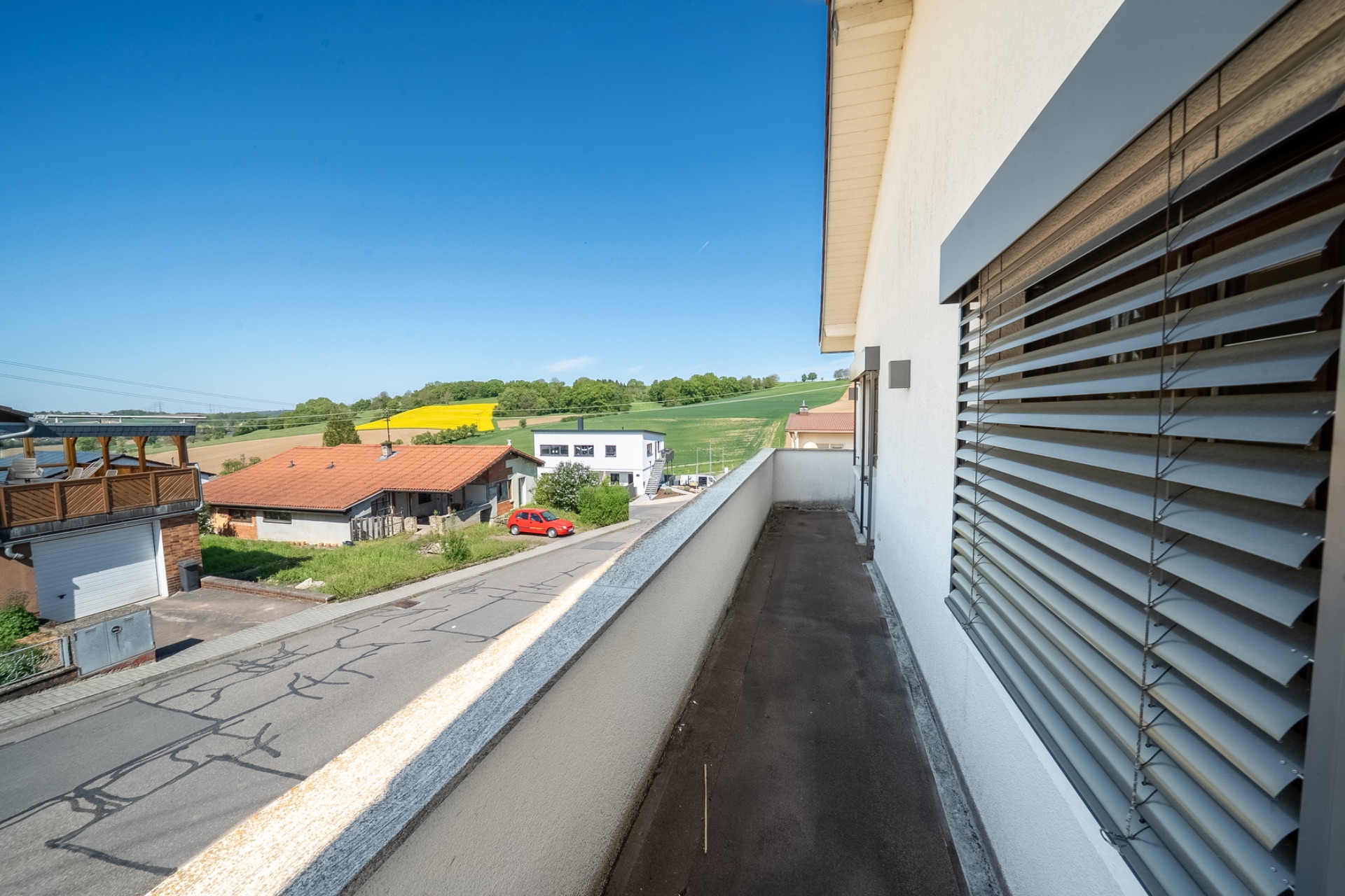 Schmaler Balkon mit Blick auf Straße, Wohnhäuser und grüne Felder im Hintergrund.