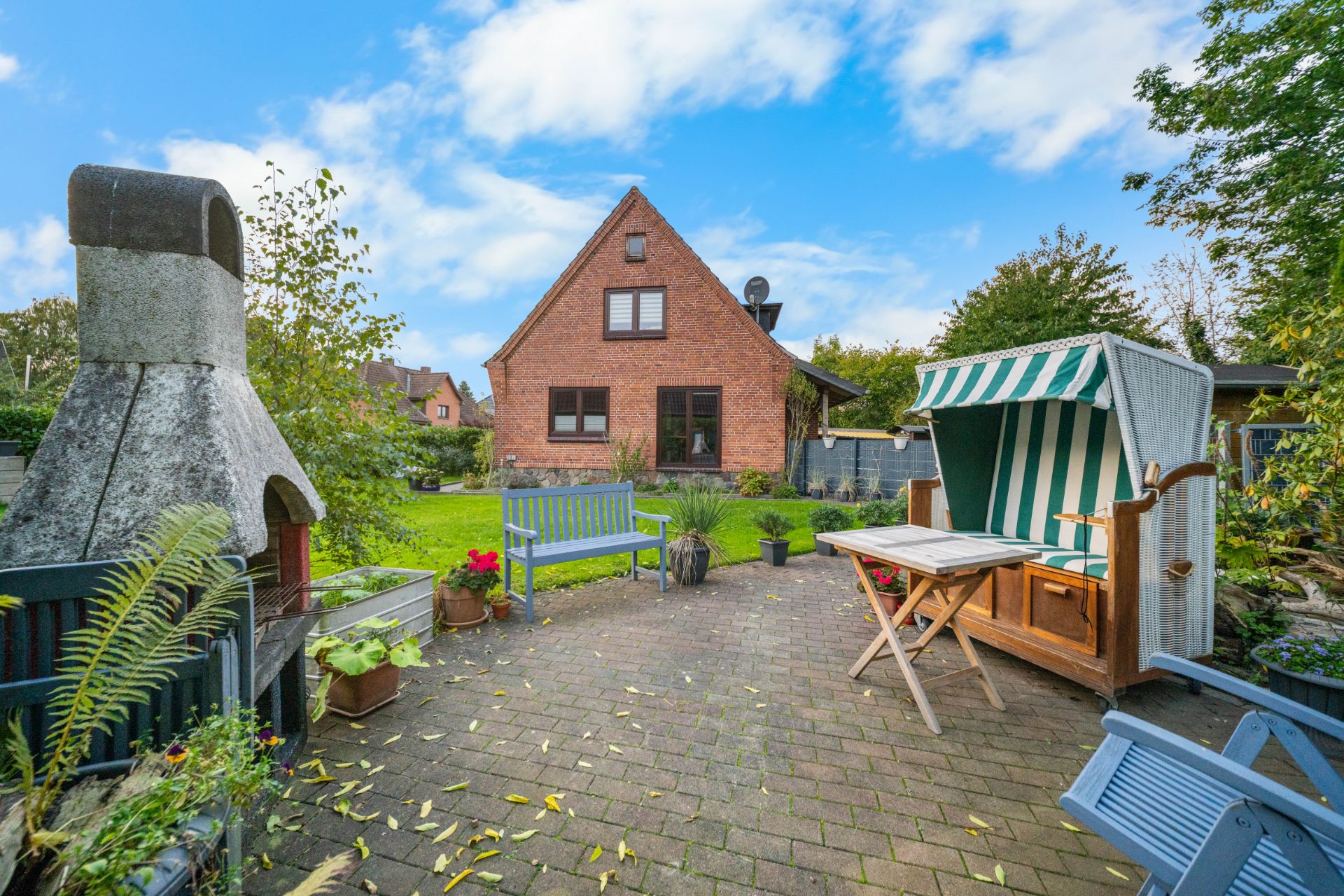 Gepflasterte Terrasse mit Sitzbänken, Strandkorb, Grill und Blick auf ein rotes Einfamilienhaus.