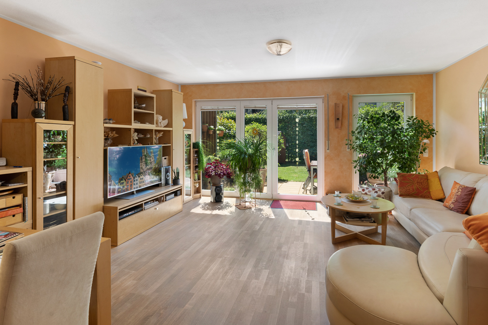 Helles Wohnzimmer mit Holzboden, Sofa, Wohnwand, vielen Pflanzen und Blick auf den Garten.