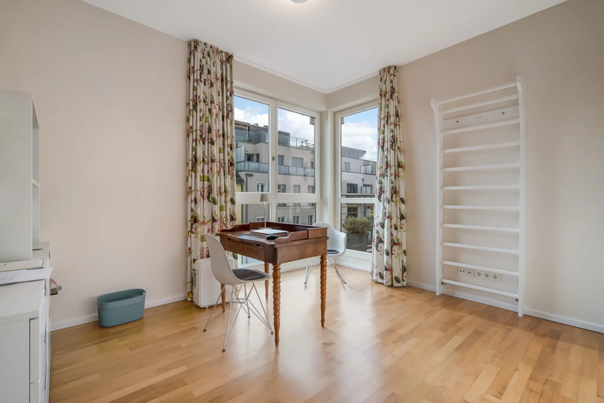 Helles Zimmer mit Holzfußboden, Schreibtisch, Stuhl, Regal und großen Fenstern mit Blumenvorhängen.