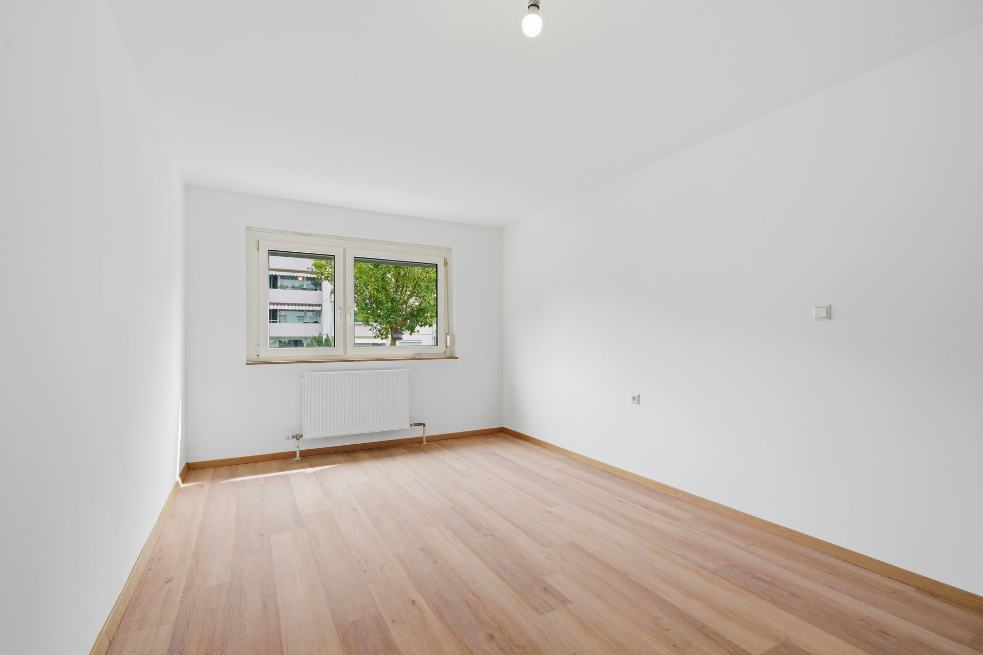 Leeres Zimmer mit Holzfußboden, weißer Wand, Fenster und Heizkörper unter dem Fenster.