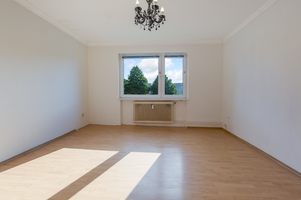 Leeres Zimmer mit Holzfußboden, zwei Fenstern, Heizkörper und Kronleuchter.