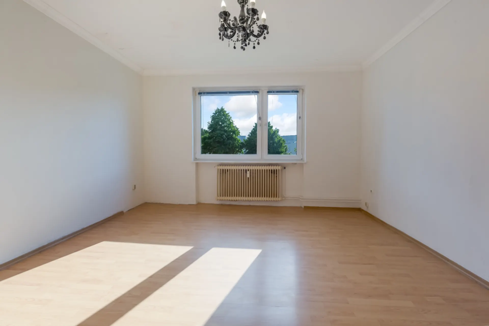 Leeres Zimmer mit Holzfußboden, zwei Fenstern, Heizkörper und Kronleuchter.