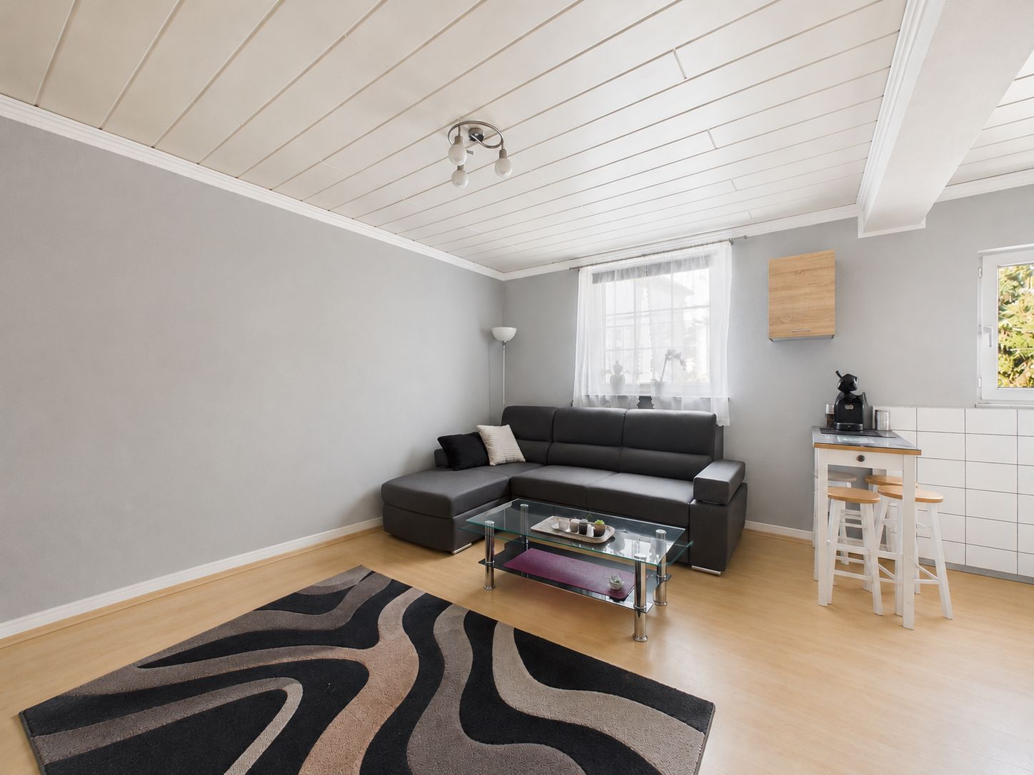Modernes Wohnzimmer mit Ecksofa, Glastisch, Teppich und kleiner Küchenecke.