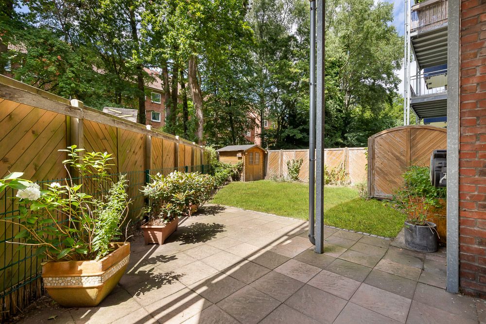 Gepflasterte Terrasse mit Pflanzen, angrenzender Rasenfläche und einem Holzhaus im Garten.