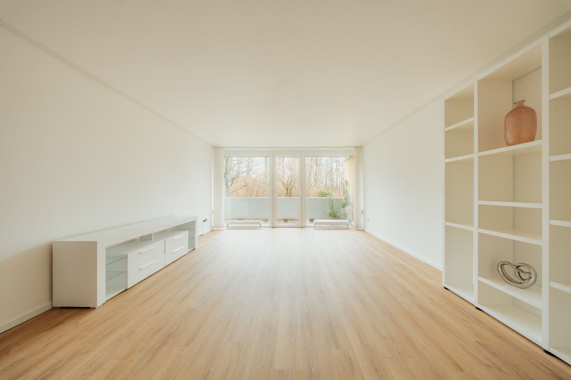 Heller, leerer Wohnraum mit Holzfußboden, Regal und großen Fenstern zur Terrasse.