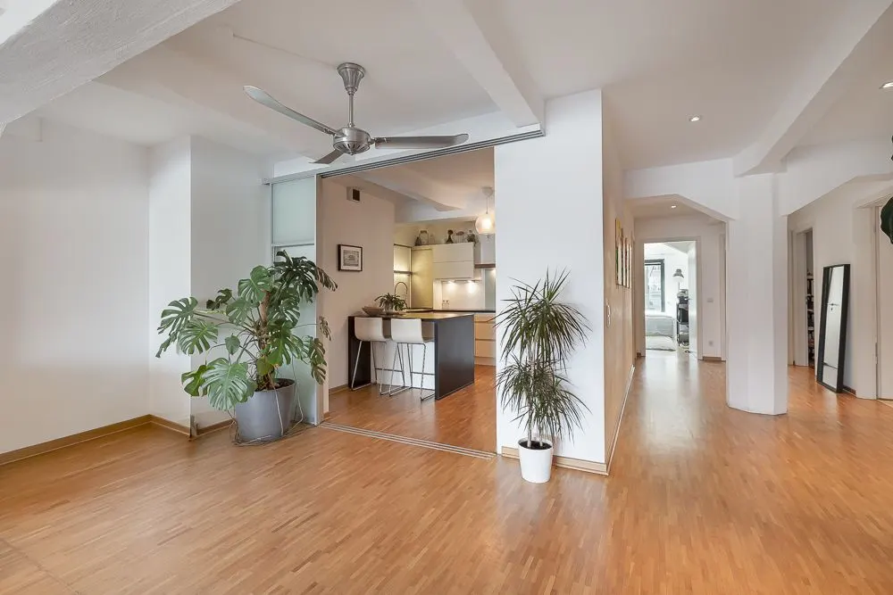 Heller Wohnraum mit Holzboden, Deckenventilator, Pflanzen und Blick in eine moderne Küche.