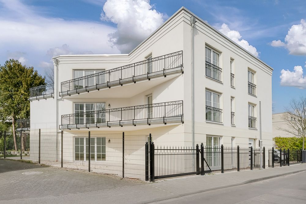 Modernes, weißes Wohngebäude mit Balkonen und schwarzem Zaun an einer Straßenecke.